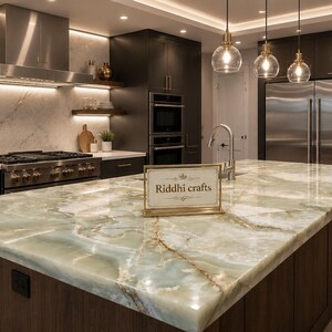 May include: A kitchen island featuring a polished light green and white marble countertop with a gold-framed sign displaying the text Riddhi crafts. The island is set in a kitchen with dark wood cabinetry, stainless steel appliances, and pendant lights.