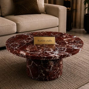 May include: A round coffee table made of deep red marble with prominent white veining. The table features a cylindrical base and a flat circular top. A rectangular gold-toned plaque with the text Riddhi crafts is centered on the tabletop.