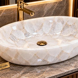 May include: An oval-shaped bathroom sink crafted from translucent white quartz stone with a polished finish. The basin features a central gold-toned drain and is positioned on a marble countertop. A small gold plaque displays the text Riddhi crafts.