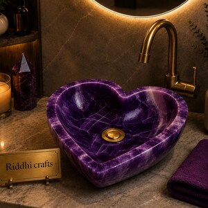 May include: A heart-shaped purple amethyst sink with a gold drain and faucet. The sink is made of polished stone and has a unique veining pattern. A small sign reads "Riddhi crafts".