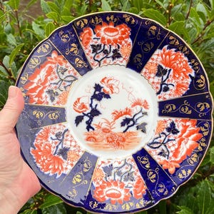 May include: A decorative plate with a blue rim and a white centre. The plate features orange and blue floral designs and a gold trim. The plate is held by a person.