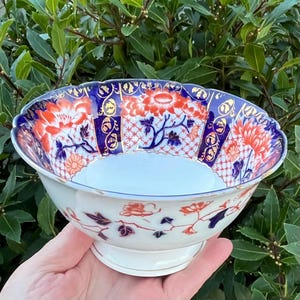 May include: A white porcelain bowl with a scalloped rim, decorated with a floral pattern in orange, blue, and gold. The bowl has a pedestal base and is set against green foliage. The floral design features stylized flowers and detailed patterns.