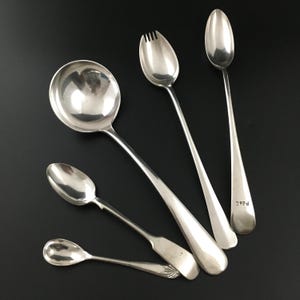 Peut inclure: Cinq cuillères en argent, dont une cuillère de service, une cuillère à fourchette et trois cuillères plus petites. La cuillère de service a un bol rond et un long manche. La cuillère à fourchette a une petite fourchette au bout du bol. Les cuillères plus petites ont des bols ronds et des manches courts. Les cuillères sont toutes polies et brillantes.