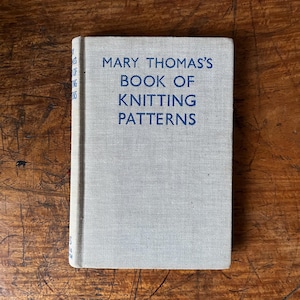 May include: A vintage book titled "MARY THOMAS'S BOOK OF KNITTING PATTERNS" in blue text on a light grey cover. The book is set against a wooden surface, showcasing its aged appearance and classic design. The book's spine is visible.