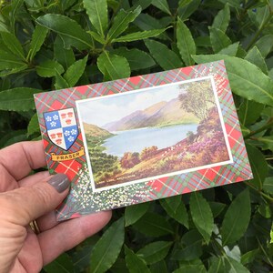 May include: A vintage postcard depicting a scenic view of Loch Long, Argyllshire, Scotland. The postcard has a red, green and blue tartan border with a crest and the word "Fraser" printed on it.