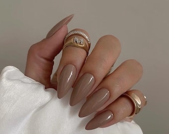 May include: A hand with long, almond-shaped, taupe-colored nails. The hand is adorned with gold rings, including one with a diamond-like design and another with the letter "M". The nails have a glossy finish.