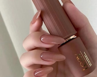 May include: A close-up of a hand with long, oval-shaped nails painted with a light brown nail polish. The hand is holding a bottle of nail polish with a gold cap and the word "LOCA" printed on the label.