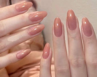 Puede incluir: Primer plano de manos con uñas largas en forma de almendra. Las uñas están pintadas con un esmalte brillante de color melocotón claro. El esmalte tiene un brillo sutil, que refleja la luz. El efecto general es elegante y pulido.