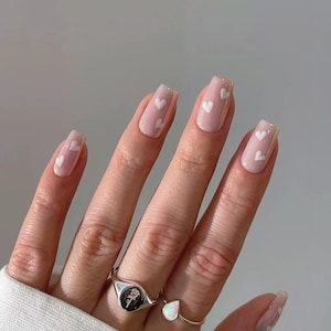 Pode incluir: Close-up de uma mão com unhas quadradas de cor rosa claro com desenhos de corações brancos. A mão está usando dois anéis de prata, um com um design de rosa e o outro com uma pedra em forma de lágrima.