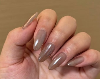 Puede incluir: Una mano con uñas largas, pulidas y en forma de almendra. Las uñas están pintadas de un color marrón neutro brillante, que refleja la luz. La manicura es de un estilo sencillo y elegante.