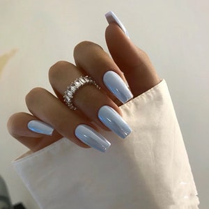 May include: Close-up of a hand with long, light blue, coffin-shaped nails. The nails are polished with a glossy finish. The hand is wearing a silver ring with a row of clear, rectangular stones.
