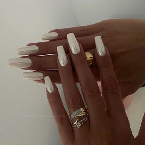 Peut inclure: Gros plan sur des mains avec de longs ongles blancs nacrés. Les ongles sont en forme de cercueil. Les mains portent des bagues en or avec de petits diamants.