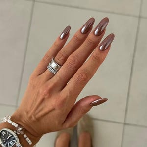 May include: A hand displaying long, almond-shaped nails in a metallic brown hue. A silver ring with multiple bands and a watch with a silver strap and black face are also visible. The nails have a glossy finish.