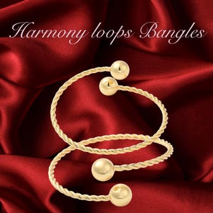 May include: Three gold-toned bangle bracelets with twisted rope designs and round ball accents. The bracelets are stacked on top of each other on a red satin background. The text "Harmony loops Bangles" is displayed above the bracelets.