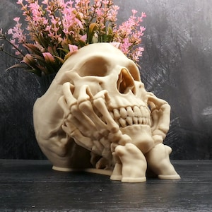 May include: A beige skull-shaped planter with hands supporting the chin. The skull has detailed teeth and eye sockets. Pink and green flowers are arranged in the skull's head. The planter is on a dark surface.