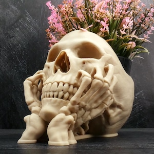 Peut inclure: Un pot de fleurs en forme de cr&acirc;ne beige avec un motif de main squelettique reposant contre la m&acirc;choire. Le cr&acirc;ne pr&eacute;sente des orbites et une cavit&eacute; nasale creuses, avec un bouquet de fleurs artificielles roses et vertes sur le dessus.