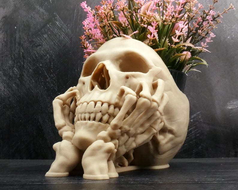 Peut inclure: Un pot de fleurs en forme de cr&acirc;ne beige avec un motif de main squelettique reposant contre la m&acirc;choire. Le cr&acirc;ne pr&eacute;sente des orbites et une cavit&eacute; nasale creuses, avec un bouquet de fleurs artificielles roses et vertes sur le dessus.