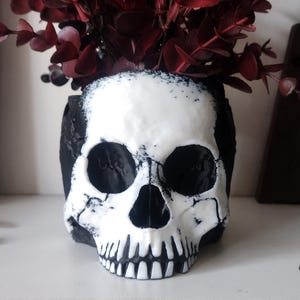 May include: A skull-shaped planter featuring a stark white face with black detailing around the eye sockets, nasal cavity, and teeth. The sides and back of the skull are solid black. Dark red eucalyptus leaves are arranged inside the top opening.