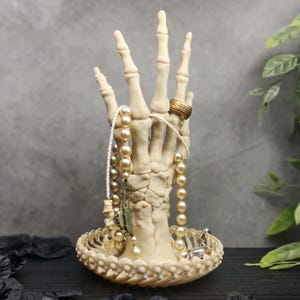 May include: A white skeletal hand ring holder with a pearl necklace and a gold ring. The hand is sitting on a white base with a scalloped edge.