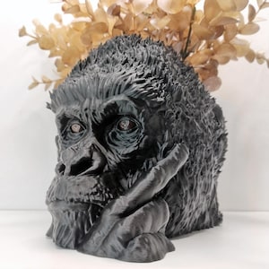 May include: A black gorilla head planter with a textured, fur-like surface and a contemplative expression. The sculpture features a hollow top opening, currently holding a sprig of dried, light brown eucalyptus leaves, creating a striking contrast.