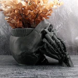 Peut inclure: Un pot de fleurs noir mat en forme de cr&acirc;ne humain avec une main squelettique reposant sur le c&ocirc;t&eacute;. Le sommet du cr&acirc;ne est ouvert, r&eacute;v&eacute;lant une texture int&eacute;rieure nervur&eacute;e, et il est rempli d'un bouquet de feuilles d'eucalyptus s&eacute;ch&eacute;es marron.
