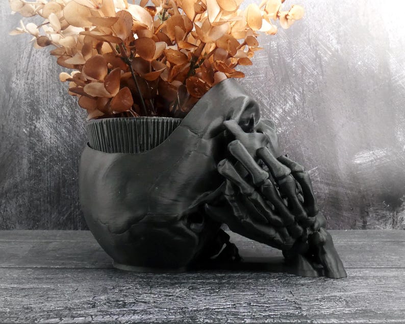 Peut inclure: Un pot de fleurs noir mat en forme de cr&acirc;ne humain avec une main squelettique reposant sur le c&ocirc;t&eacute;. Le sommet du cr&acirc;ne est ouvert, r&eacute;v&eacute;lant une texture int&eacute;rieure nervur&eacute;e, et il est rempli d'un bouquet de feuilles d'eucalyptus s&eacute;ch&eacute;es marron.