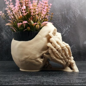 Peut inclure: Un pot de fleurs en forme de cr&acirc;ne beige avec une main squelettique pos&eacute;e sur le c&ocirc;t&eacute; du cr&acirc;ne. Le sommet du cr&acirc;ne est ouvert et contient un pot en plastique noir rempli de fleurs artificielles rose clair avec des tiges et des feuilles vertes.