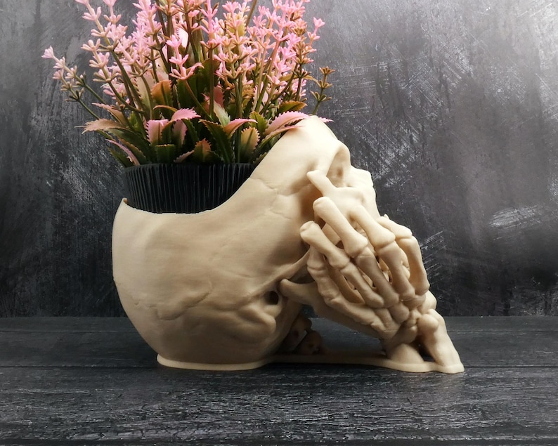 Peut inclure: Un pot de fleurs en forme de cr&acirc;ne beige avec une main squelettique pos&eacute;e sur le c&ocirc;t&eacute; du cr&acirc;ne. Le sommet du cr&acirc;ne est ouvert et contient un pot en plastique noir rempli de fleurs artificielles rose clair avec des tiges et des feuilles vertes.