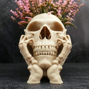 Peut inclure: Un pot de fleurs en forme de cr&acirc;ne beige avec deux mains squelettiques reposant contre les c&ocirc;t&eacute;s de la m&acirc;choire. Le sommet du cr&acirc;ne est ouvert et contient un bouquet de fleurs artificielles roses avec des tiges et des feuilles vertes.