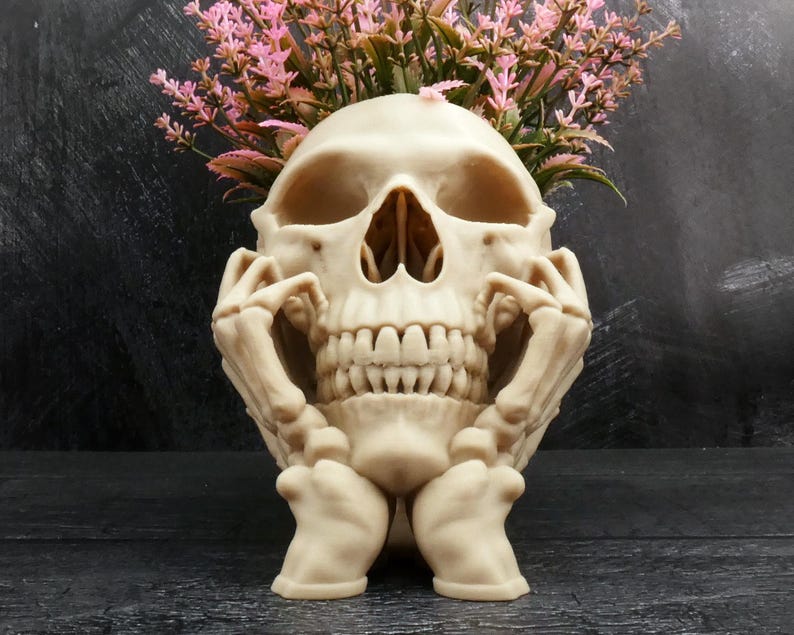Peut inclure: Un pot de fleurs en forme de cr&acirc;ne beige avec deux mains squelettiques reposant contre les c&ocirc;t&eacute;s de la m&acirc;choire. Le sommet du cr&acirc;ne est ouvert et contient un bouquet de fleurs artificielles roses avec des tiges et des feuilles vertes.