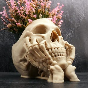 Peut inclure: Un pot de fleurs en forme de cr&acirc;ne beige avec une main squelettique reposant contre le menton et la m&acirc;choire. Le cr&acirc;ne poss&egrave;de un sommet creux contenant un bouquet de fleurs artificielles roses aux tiges vertes. Finition mate couleur os.