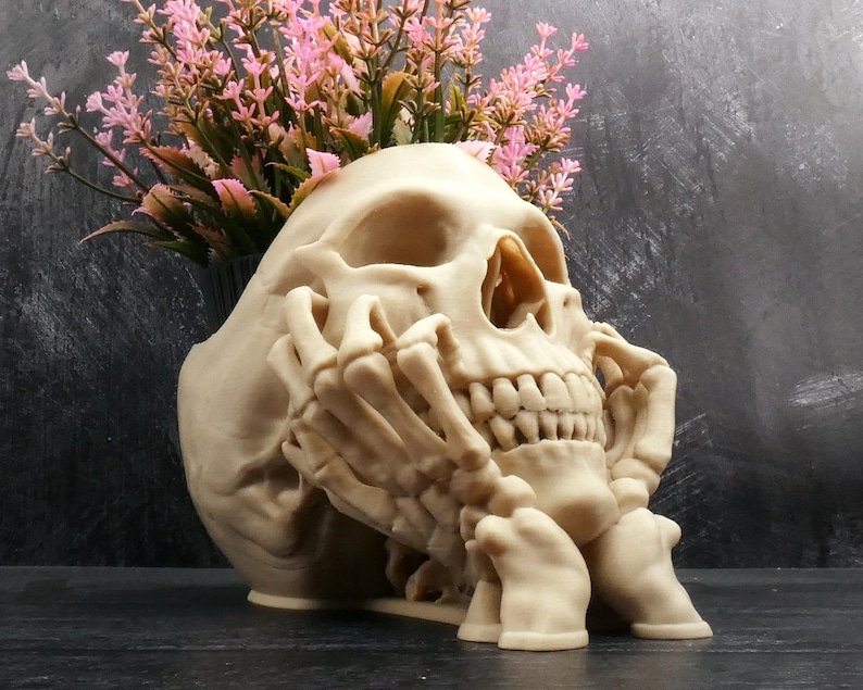 Peut inclure: Un pot de fleurs en forme de cr&acirc;ne beige avec une main squelettique reposant contre le menton et la m&acirc;choire. Le cr&acirc;ne poss&egrave;de un sommet creux contenant un bouquet de fleurs artificielles roses aux tiges vertes. Finition mate couleur os.