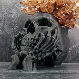 Peut inclure: Un pot de fleurs en forme de cr&acirc;ne noir mat avec un motif de main squelettique soutenant le menton. Le cr&acirc;ne pr&eacute;sente des orbites profondes et une m&acirc;choire d&eacute;taill&eacute;e, avec un bouquet de feuilles d'eucalyptus s&eacute;ch&eacute;es brun clair au sommet.