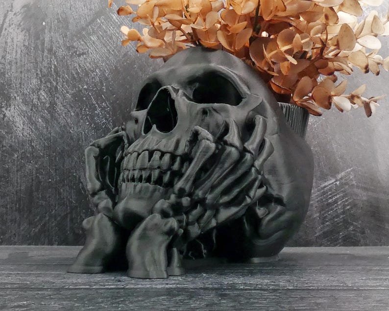 Peut inclure: Un pot de fleurs en forme de cr&acirc;ne noir mat avec un motif de main squelettique soutenant le menton. Le cr&acirc;ne pr&eacute;sente des orbites profondes et une m&acirc;choire d&eacute;taill&eacute;e, avec un bouquet de feuilles d'eucalyptus s&eacute;ch&eacute;es brun clair au sommet.
