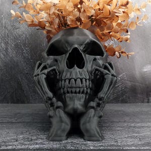 Peut inclure: Un pot de fleurs en forme de cr&acirc;ne noir mat avec des mains squelettiques reposant sur les c&ocirc;t&eacute;s du visage. Le cr&acirc;ne creux contient un bouquet de branches d'eucalyptus s&eacute;ch&eacute;es brun clair, cr&eacute;ant un contraste saisissant avec la finition sombre.