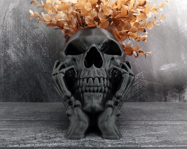Peut inclure: Un pot de fleurs en forme de cr&acirc;ne noir mat avec des mains squelettiques reposant sur les c&ocirc;t&eacute;s du visage. Le cr&acirc;ne creux contient un bouquet de branches d'eucalyptus s&eacute;ch&eacute;es brun clair, cr&eacute;ant un contraste saisissant avec la finition sombre.