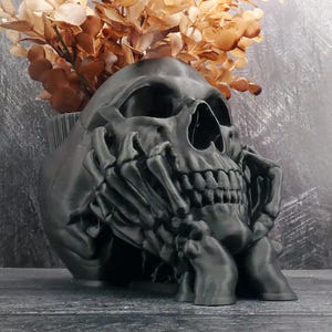 Peut inclure: Un pot de fleurs en forme de cr&acirc;ne noir mat avec un motif de mains squelettiques soutenant le menton et la m&acirc;choire. Le cr&acirc;ne creux sert de r&eacute;cipient et contient un bouquet de feuilles et de tiges s&eacute;ch&eacute;es de couleur brun clair.