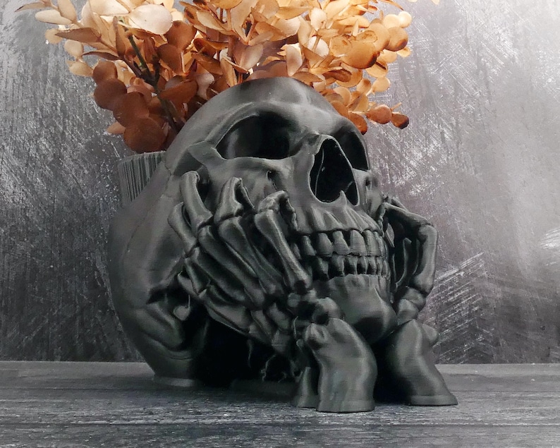 Peut inclure: Un pot de fleurs en forme de cr&acirc;ne noir mat avec un motif de mains squelettiques soutenant le menton et la m&acirc;choire. Le cr&acirc;ne creux sert de r&eacute;cipient et contient un bouquet de feuilles et de tiges s&eacute;ch&eacute;es de couleur brun clair.