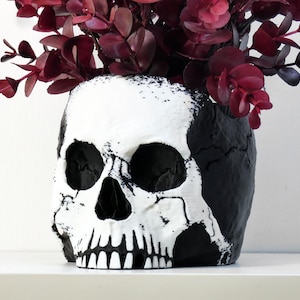 May include: A skull-shaped planter featuring a high-contrast design with a matte black base and a textured white face. The hollow cranium is filled with a cluster of deep burgundy eucalyptus branches, creating a striking visual display of dark foliage.