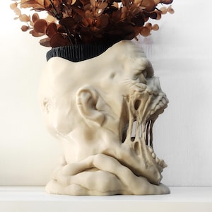 May include: A beige, sculpted planter shaped like a decaying human head with an open, screaming mouth and exposed teeth. The top of the head features a black circular opening containing a small, brown, leafy plant with multiple stems.