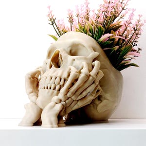 May include: A beige skull-shaped planter with hands cradling the face, filled with pink and green artificial flowers. The skull has detailed teeth and eye sockets, creating a unique decorative piece for home decor.