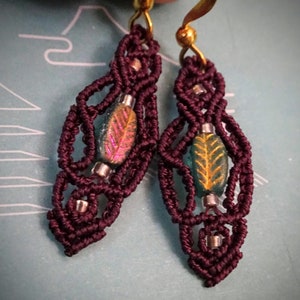 May include: A pair of burgundy macrame earrings with gold hooks and iridescent green and pink leaf-shaped beads.