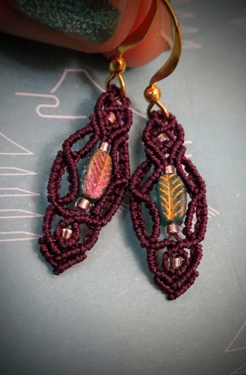 May include: A pair of burgundy macrame earrings with gold hooks and iridescent green and pink leaf-shaped beads.