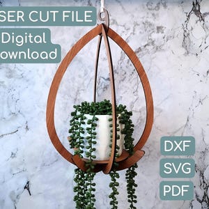 May include: A wooden hanging planter with a white ceramic pot filled with greenery. The planter has a teardrop-shaped design and includes the text "LASER CUT FILE Digital Download". The image also shows the file types: DXF, SVG, and PDF.