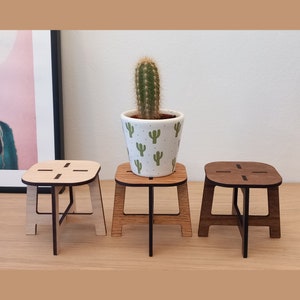 May include: Three wooden plant stands, each with a different wood stain. The stand in the center has a small cactus plant in a white ceramic pot with a cactus pattern.