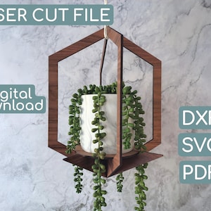 Pode incluir: Um vaso de plantas suspenso de madeira com um design geométrico, segurando um vaso branco e cinza marmorizado cheio de plantas verdes penduradas. As palavras "LASER CUT FILE" e "Digital Download" são visíveis. Inclui formatos de arquivo DXF, SVG e PDF.