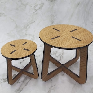 May include: Two small, wooden stools with a natural wood grain finish. Each stool has a round top with four cut-out slots and an X-shaped base. The stools are different sizes, with the larger one on the right.