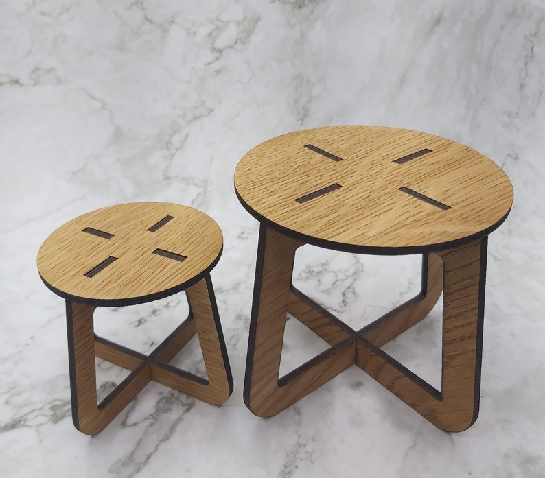 May include: Two small, wooden stools with a natural wood grain finish. Each stool has a round top with four cut-out slots and an X-shaped base. The stools are different sizes, with the larger one on the right.