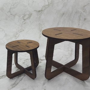 May include: Two wooden side tables. The larger table has a round top with four cut-out slots and an X-shaped base. The smaller table has a similar design. Both tables are made of dark brown wood and are set against a white marble background.