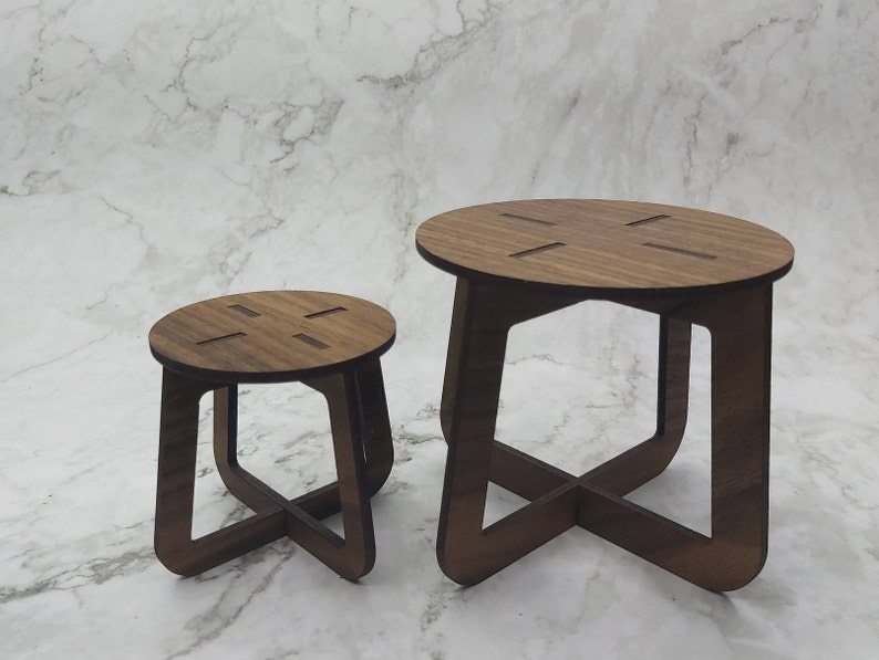 May include: Two wooden side tables. The larger table has a round top with four cut-out slots and an X-shaped base. The smaller table has a similar design. Both tables are made of dark brown wood and are set against a white marble background.